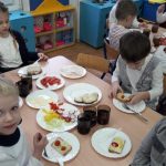 Kilkoro dzieci siedzi na krześle przy stole. Na stole leżą talerze, pokrojone warzywa, chleb z masłem i kubek z herbatą.