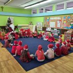 Dzieci siedzią wraz z Mikołajem w kole na dywanie. Na głowach mają czapki mikołajkowe.