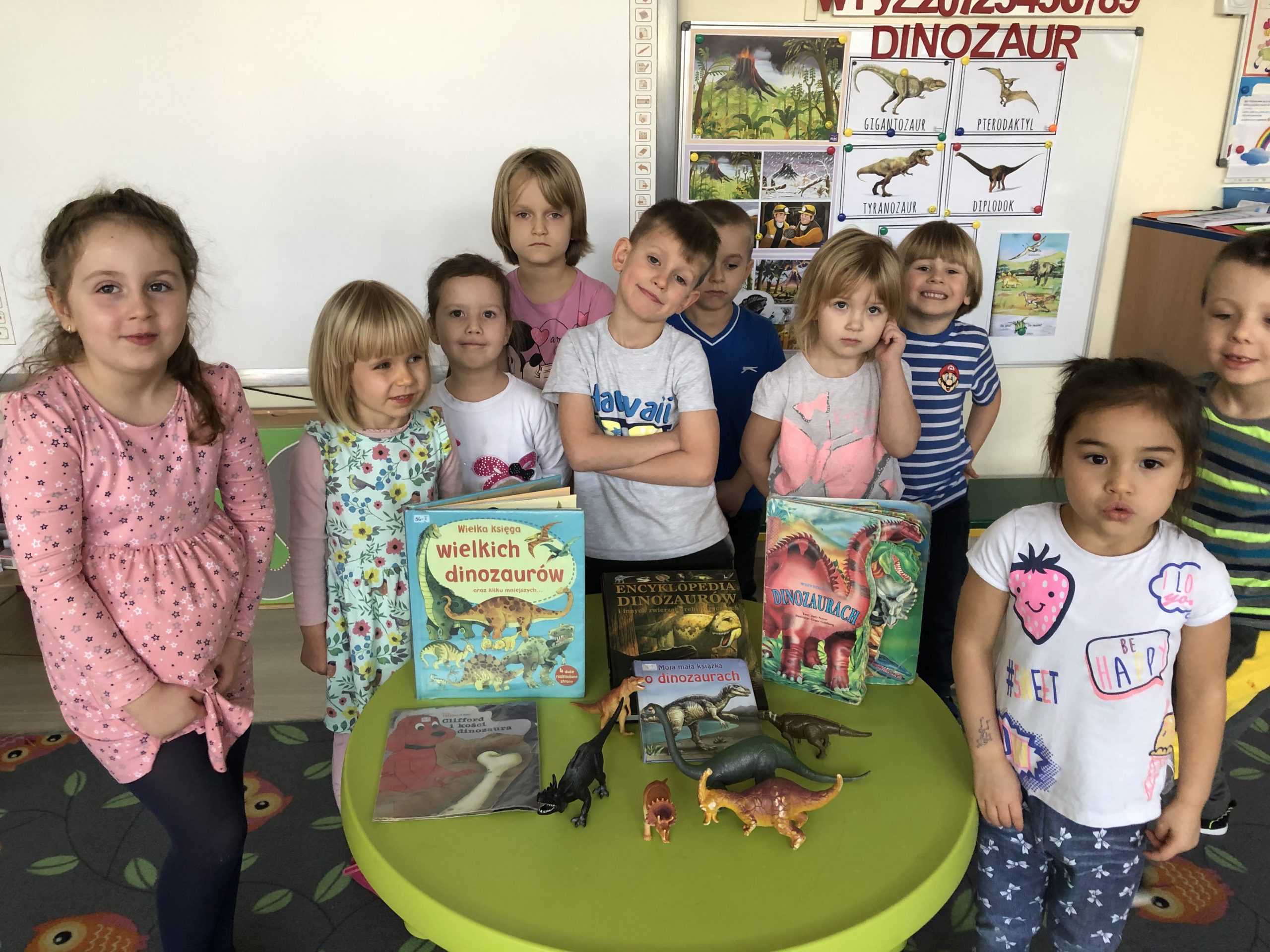 Dzieci stojący w grupie. Przed nimi na stoliku znajdują się książki o tematyce dinozaurów oraz figurki dinozaurów.