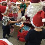 Kilkoro dzieci stojących na dywanie. Z prawej strony Mikołaj podaje rękę chłopcu. Przed Mikołajem znajduje się worek z prezentami.