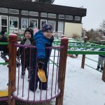 Kilkoro dzieci bawiących się na pokrytym śniegiem, przedszkolnym placu zabaw. Chłopiec będący na pierwszym planie trzyma w ręku plastikową łopatkę..