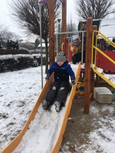 Chłopiec zjeżdżający na pokrytej śniegiem zjeżdżalni, znajdującej się na przedszkolnym placu zabaw. Za nim kuca jeszcze jedno dziecko.