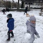 Dzieci i Panie bawiące się w pokrytym śniegiem ogrodzie przedszkolnym. Dziewczynka znajdująca się na pierwszym planie trzyma w ręku kulkę śniegu.