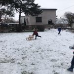 Troje dzieci bawiących się na śniegu w ogrodzie przedszkolnym. Jeden chłopiec popycha sanki.