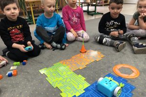 Pięcioro dzieci siedzących na dywanie w sali przedszkolnej. Jeden chłopiec trzyma w rękach pilota. .Przed dziećmi, znajduje się ułożona z puzzli trasa, oraz robot, wieżyczka, koło i plastikowe, kolorowe kostki.