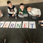 Pięcioro dzieci siedzących na dywanie i trzymających w rękach wełnę tworzącą sieć. Przed dziećmi, na dywanie znajduje się napis INTERNET.