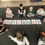 Grupa dzieci siedzących w kole na dywanie i trzymających w rękach wełnę tworzącą sieć. W środku, na podłodze znajduje się napis INTERNET.