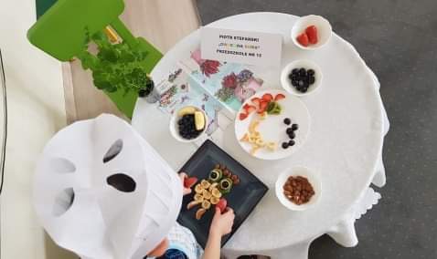 Dziecko mające na głowie czapkę kucharską siedzące przy stole, na którym znajdują się talerze i miseczki z owocami. Dziecko układa na talerzu owocową sowę.