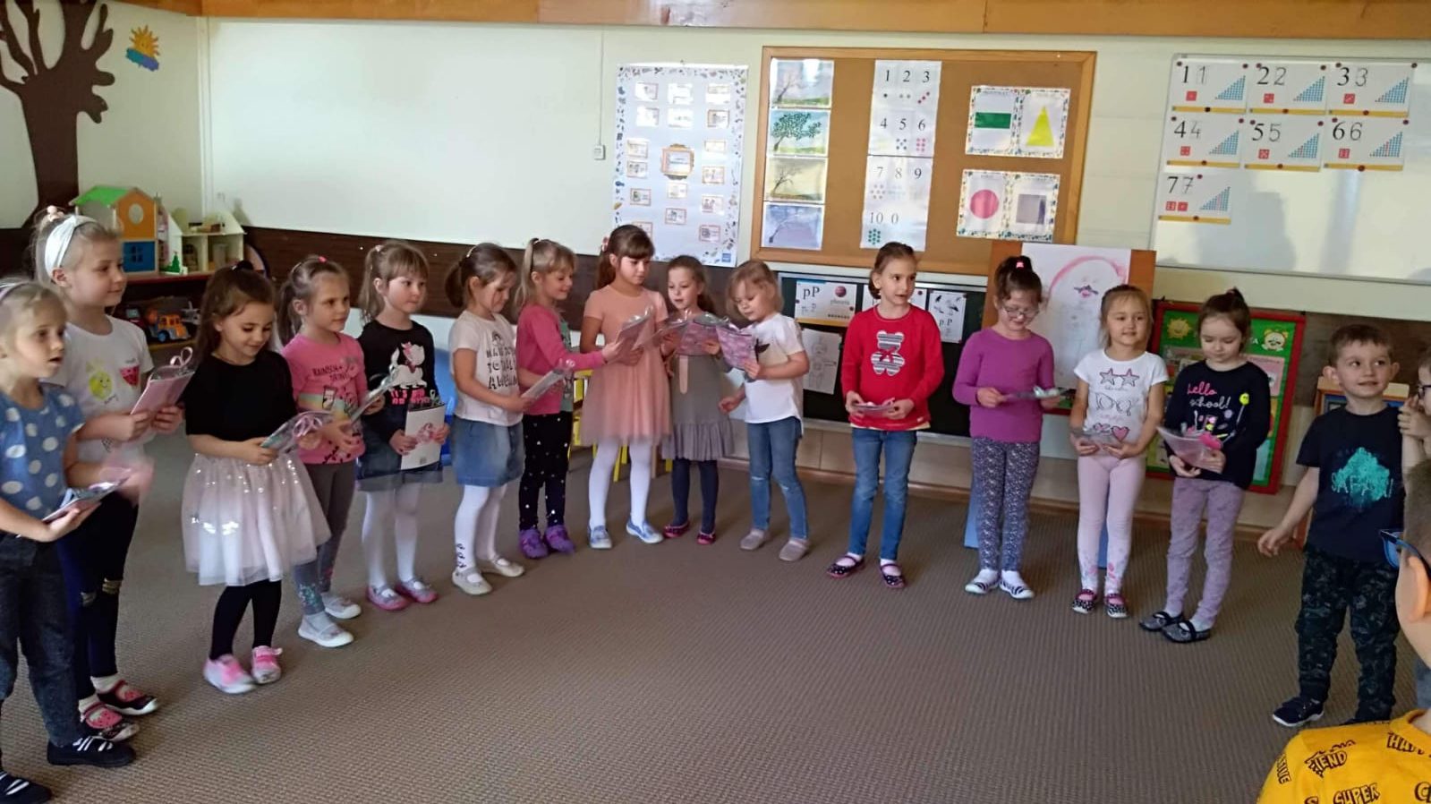 Grupa dzieci stojąca w sali przedszkolnej i trzymająca w rękach prezenty. w półkolu