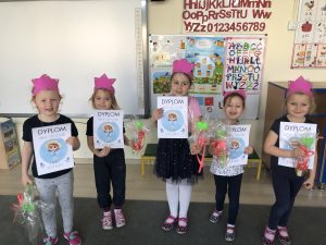 Pięć dziewczynek mających na głowach różowe opaski-korony stojące w jednym rzędzie w sali przedszkolnej. Każda dziewczynka trzyma w rękach upominek oraz dyplom.