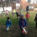 Grupa dzieci zanajdujących się w ogrodzie przedszkolnym, szukających wielkanocnych koszyczków. Troje dzieci trzyma w rękach koszyczki. Za dziećmi stoi Pani.