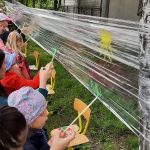 Dzieci na dworze malują farbami na folii zawieszonej pomiędzy 2 drzewami.