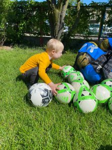 Chłopiec kucający w ogrodzie przedszkolnym i trzymajacy w rękach piłki.