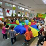 Grupa dzieci stoi w kole trzymając w rękach kolorową chustę z balonami.
