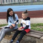 Chłopiec z nastolatką siedzący na ławce, na boisku szkolnym. Dziewczyna trzyma w ręku mikrofon, chłopiec udziela wywiadu.