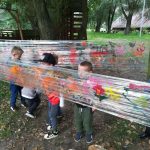 Sześcioro dzieci malujacych na dwo9rze farbami po folii rozłożonej między drzewami.