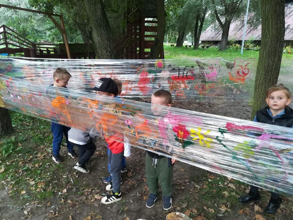 Sześcioro dzieci malujacych na dwo9rze farbami po folii rozłożonej między drzewami.