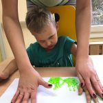 Chłopiec siedzi przy stole ma pomalowane dłonie na zielono, które odciska na kartce papieru.