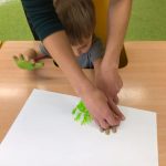 Chłopiec siedzi przy stole ma pomalowane dłonie na zielono, które odciska na kartce papieru.