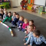 Dzieci siedzą na dywanie jeden za drugim. Obok nich leży wąż ułożony z kolorowych kubeczków. Chłopiec w zielonej bluzie trzyma na nim prawą rękę.