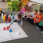 Na dywanie znajduje się kwadratowa mata, na której chłopiec układa kolorowe kubki według określonego wzoru. Obok siedzą dzieci i obserwują co robi kolega.
