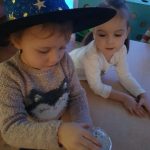 2 dziewczynki stoją przy stoliku. Jedna ma na głowie kapelusz czarodziejki.