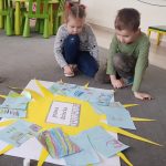 Dziewczynka i chłopiec siedzą na szarym dywanie w przedszkolnej sali. Przed chłopcami leży arkusz białego papieru, a na nim naklejone żółte słońce. Na jego promieniach są kartki z rysunkami.