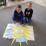 Dwóch chłopców siedzi na szarym dywanie w przedszkolnej sali. Przed chłopcami leży arkusz białego papieru, a na nim naklejone żółte słońce. Na jego promieniach są kartki z rysunkami.