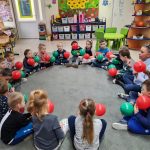 grupa dzieci siedzi w przedszkolnej sali na dywanie w kole. dzieci trzymają w rękach zielone i czerwone balony.