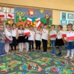Grupa 13 dzieci ustawiona w półkolu. Dzieci ubrane na galowo, w dłoni trzymają flagę Polski.
