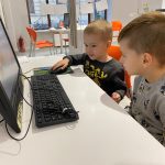 Dwóch chłopców siedzących przy biurku, patrzacych w monitor. Jeden chłopiec trzyma dłoń na myszce od komputera.