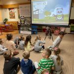 Dzieci siedzą na dywanie przed ekranem i oglądają prezentację.