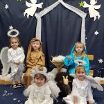 5 dzieci siedzi na dywanie lub krześle. Na środku stoi żłobek po bokach dziewczynka w przebraniu Maryi i chłopiec w przebraniu Józefa. Przed nimi siedzą dziewczynki przebrane za aniołki.