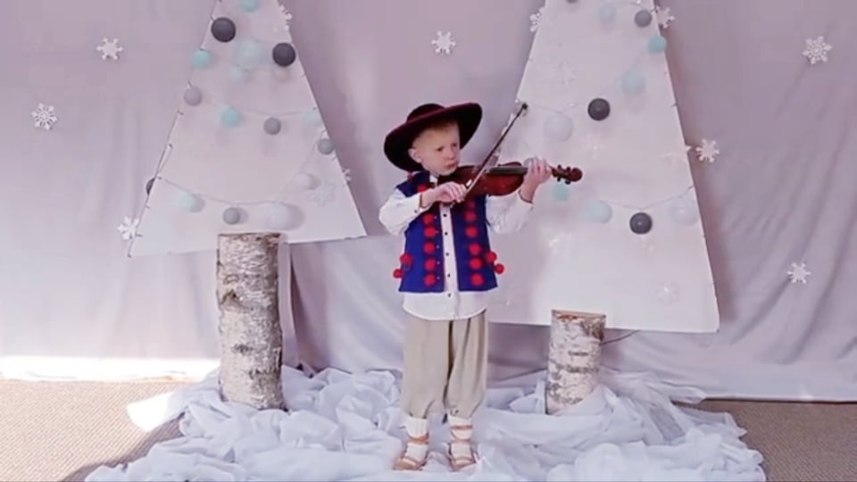 Chłopiec ubrany w strój góralski, niebieską kamizelkę, białą koszulę, jasne spodnie i kierpce grana skrzypcach. W tle stoją dwie białe choinki na pniach