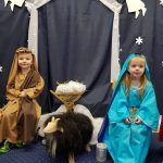 Dziewczynka przebrana za Maryję i chłopiec za Józefa siedzą na krzesłach, pośrodku stoi żłóbek i drewniania owca.