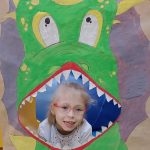 Dziewczynka siedzi na plakatem przedstawiającym dinozaura z otwartą buzią.