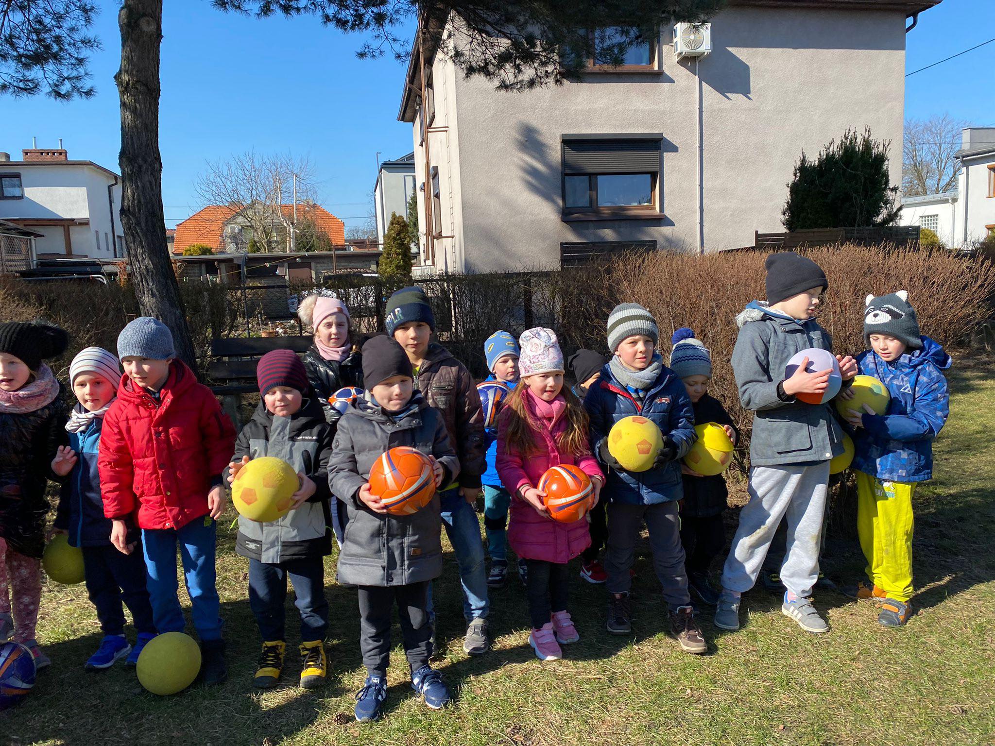 Duża grupa dzieci na dworze stoi z piłkami w ręce.