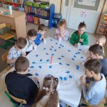 Grupa dzieci siedzi przy jednym stoliku. Każde dziecko ma przed sobą biały kubeczek z wodą i kolorowe słomki.