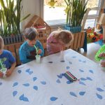 Grupa dzieci siedzi przy jednym stoliku. Każde dziecko ma przed sobą biały kubeczek z wodą i kolorowe słomki.