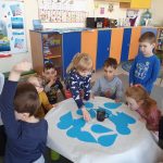 Grupa dzieci siedzi na dywanie. Na środku stoi stolik, a na nim błękitne krople wody., szklanka z wodą i podstawek.