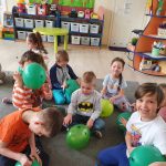 Grupa dzieci siedzi na dywanie z zielonymi balonami.