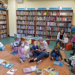Dzieci i Panie siedzą na dywanie w bibliotece. Każde dziecko czyta książeczkę.