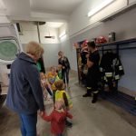 Dzieci i Panie patrzą na ana strażaka który prezentuje hełm strażacki