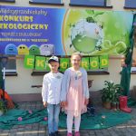 Dziewczynka i chłopiec stoją przed przedszkolem na tle plakatu , promującego konkurs.