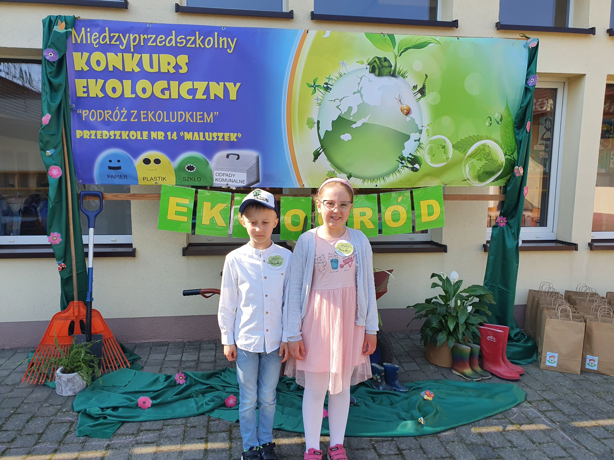 Dziewczynka i chłopiec stoją przed przedszkolem na tle plakatu , promującego konkurs.