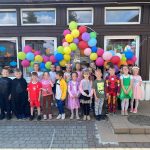 Grupa dzieci stojąca przed budynkiem przedszkola. Nad głowami wiszą kolorowe balony.