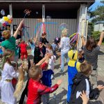 Grupa dzieci uczestnicząca w grach i zabawach ruchowych przed budynkiem przedszkola. W tle cztery Panie puszczają bańki. Na budynku przedszkola widnieją dekoracje z balonów.