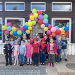 Grupa dzieci stojąca przed budynkiem przedszkola. Nad głowami wiszą kolorowe balony.