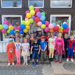 Grupa dzieci stojąca przed budynkiem przedszkola . Nad dziećmi wiszą kolorowe balony.