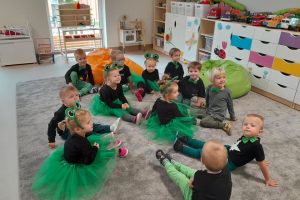Dwanaścioro dzieci siedzących na dywanie w sali przedszkolnej. Dziewczynki mają ubrane stroje żabek, chłopcy mają założone zielone muchy.